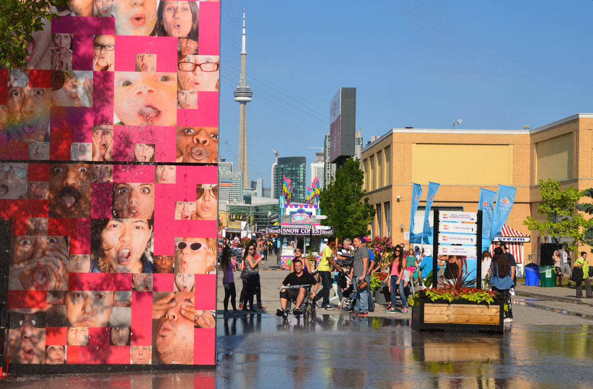 people are congregating around the base of a large water feature at the CNE, a large pink box with pictures of faces on it, the water spouts out of the mouths of the people  