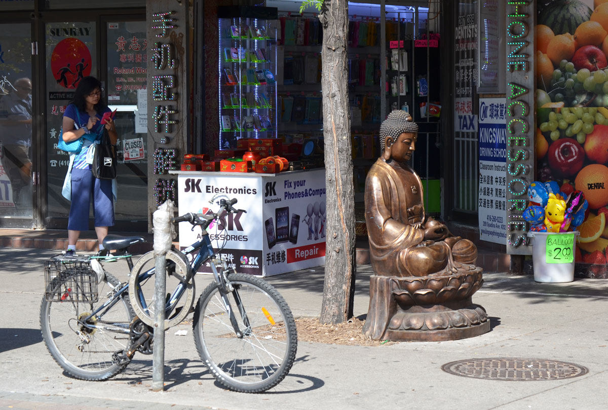 a large brown statue of a seated buddha is on the sidewalk outside a store.  A bike is parked near it and a woman is walking past. 