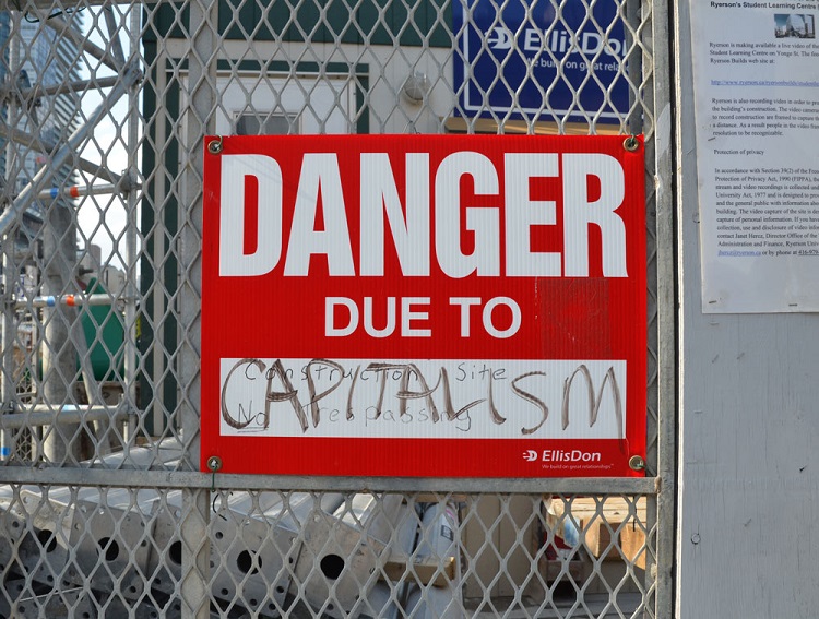 red and white construction sign that has been altered to say danger due to capitalism