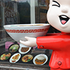 a large statue of a girl with a large white head, is winking at passersby.  She is wearing a red outfit and is holding a bowl in her hands.  She is in the window of a restaurant.