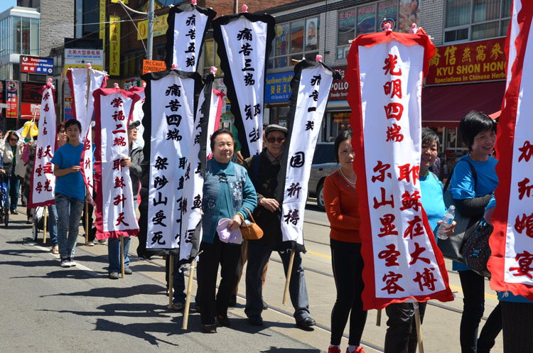 a group of Chinese people in a small parade.  They are all holding white banners with either red or blue Chinese characters on them. 