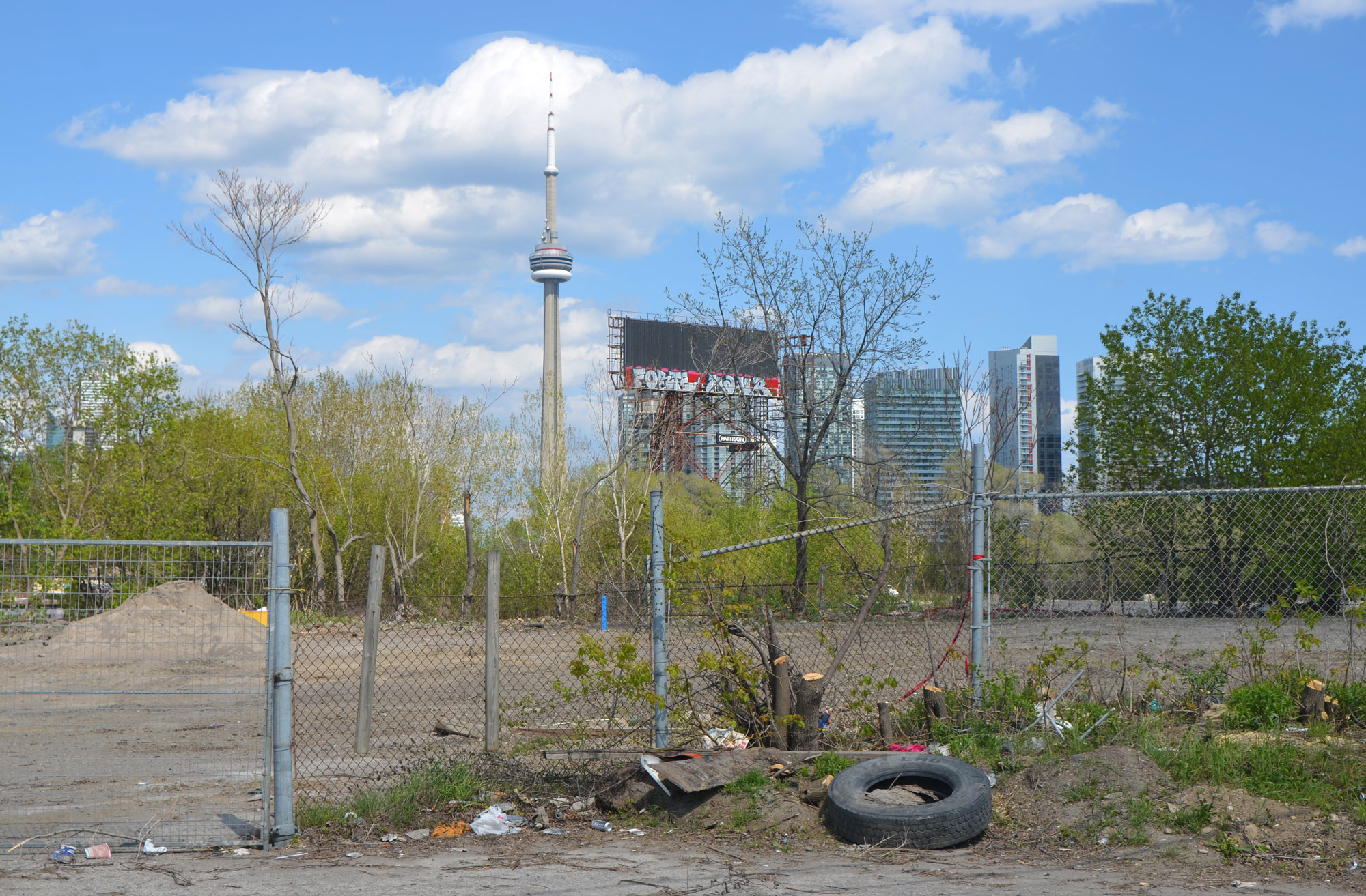 the CN Tower is in the background, a vacant lot waiting for development is in the foreground.  Old tire and some other junk is in front of the fence surrounding the vacant lot 