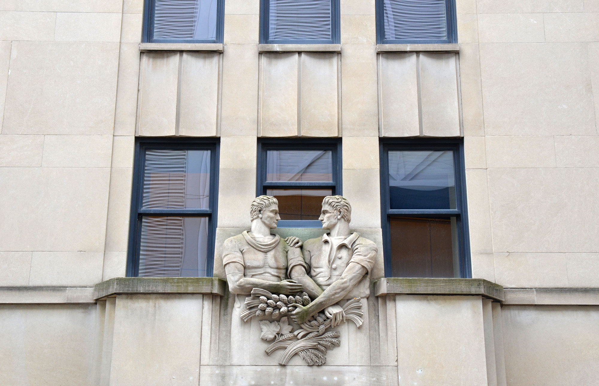 a  sculpture over the door way of a stone building, the Chang School at Ryerson University, of two men from waist up, standing beside each other with their arms entwined. They are hold stalks of wheat and other farm products  