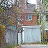 a Toronto alley, garbage bins, garages, the back of two houses, one very crooked, the back of a low rise apartment building, chainlink fence, fall foilage. 