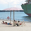 a large ship, or lake freighter, is parked in the harbour beside sugar beach where people are sitting under pink umbrellas.