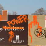 orange spray painted words, rich, power, on a fence around a construction site 