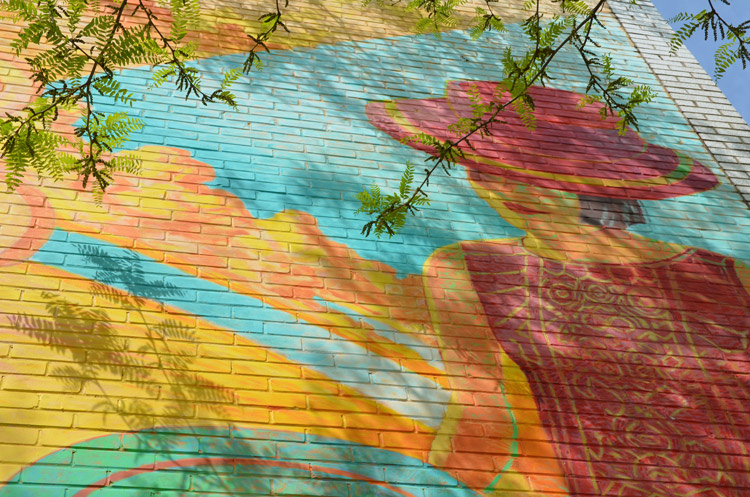 part of a mural on the side of a brick building, showing a woman who is wearing a wide brimmed pink hat