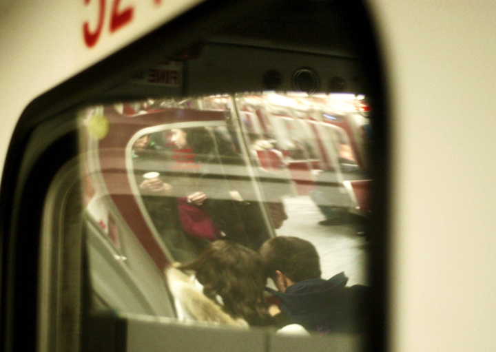 a view of the interior of a subway train.  People sitting, people standing reflected in a mirror, holding coffee cups and talking