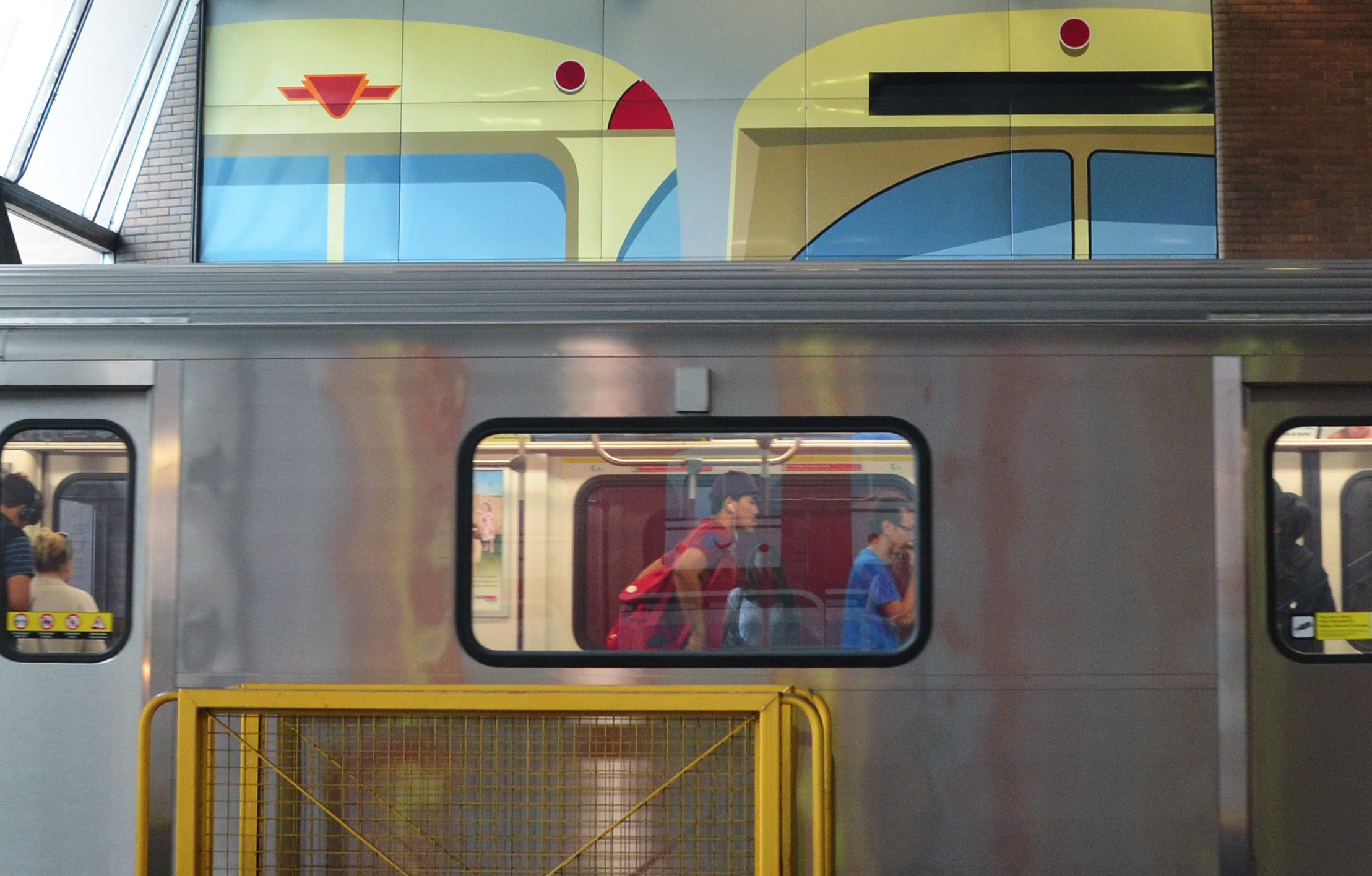 view of a subway car from the side with people in the window.  The subway is in Eglinton West station and part of the artwork by Gerald Zeldin, stylized pictures of subway cars, is on the wall behind the real subway car 