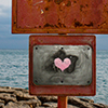 a rusty old sign on the waterfront that someone has painted a pink heart on.  Lake Ontario is in the background 