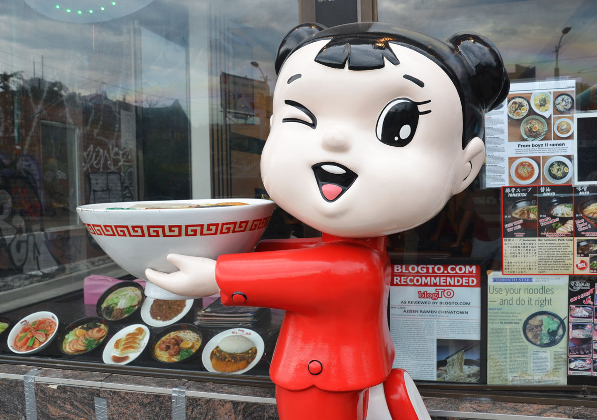 a large statue of a girl with a large white head, is winking at passersby.  She is wearing a red outfit and is holding a bowl in her hands.  She is in the window of a restaurant.
